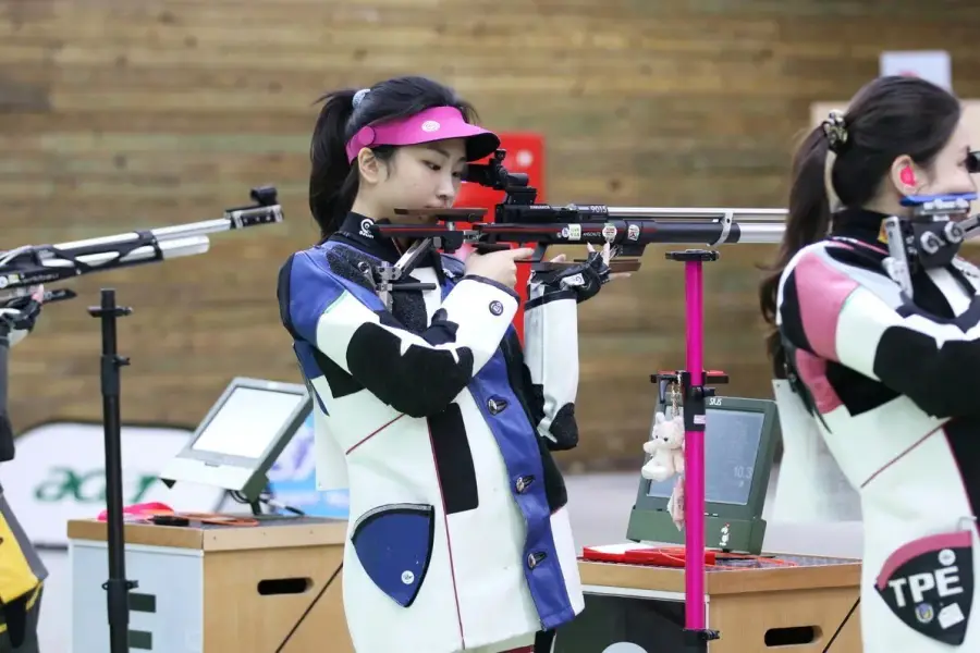 一位年輕女性選手身穿射擊夾克，在室內靶場舉起10公尺空氣步槍瞄準，背景可見比賽用的靶道標線