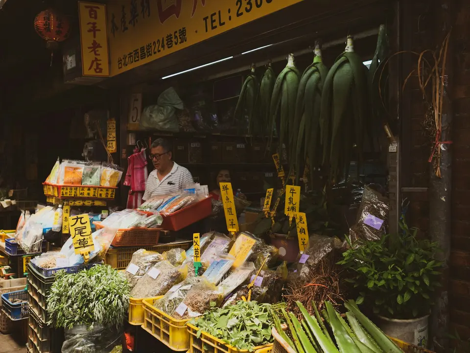 台北中藥材老店實景，男性店主站在擺滿各式乾燥藥材的攤位前，掛有中文招牌