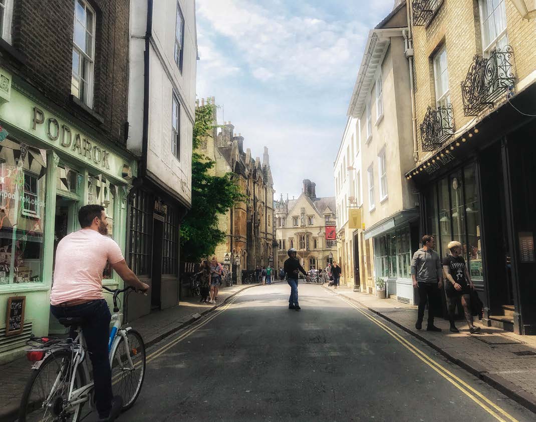 英國劍橋街道兩側的歷史建築與商店,前景有牽單車的人,街上可見行人。