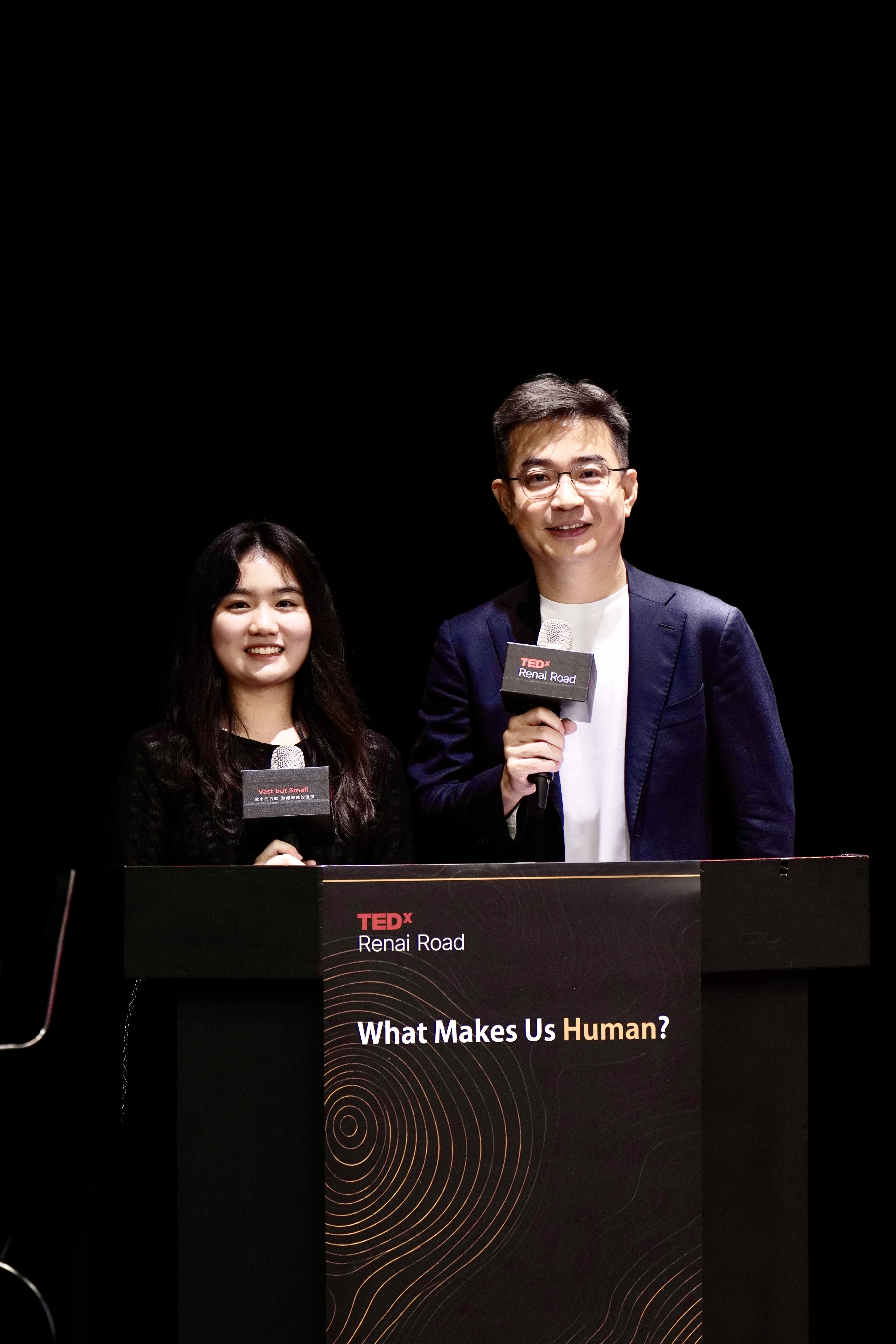 TEDxRenaiRoad主持人站在舞台講台後，畫面呈現主題文字與現場主持工作