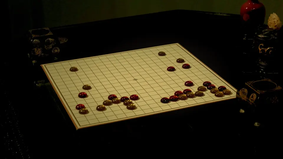 暗室木桌上擺放著白色格線圍棋棋盤，棋盤上散落著紅褐色棋子，兩側各有一個精緻黑色棋盒