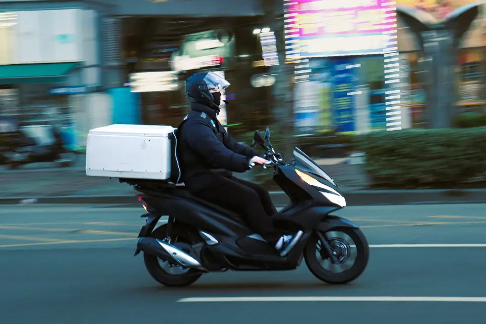 外送員騎黑色機車在城市街道疾馳，背後背著大型白色保溫外送箱