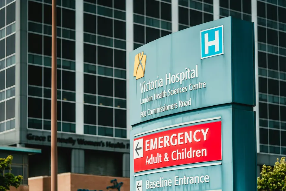 醫院門口的急診指示牌，藍色立柱上標示 Victoria Hospital 與紅底白字的 EMERGENCY Adult & Children 方向箭頭，背景為現代化玻璃帷幕大樓