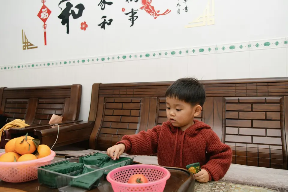 一個穿紅色針織外套的亞洲小男孩坐在木桌前，專注地撥弄桌上的分格盒子，旁邊擺著橘子，牆上有中文書法裝飾